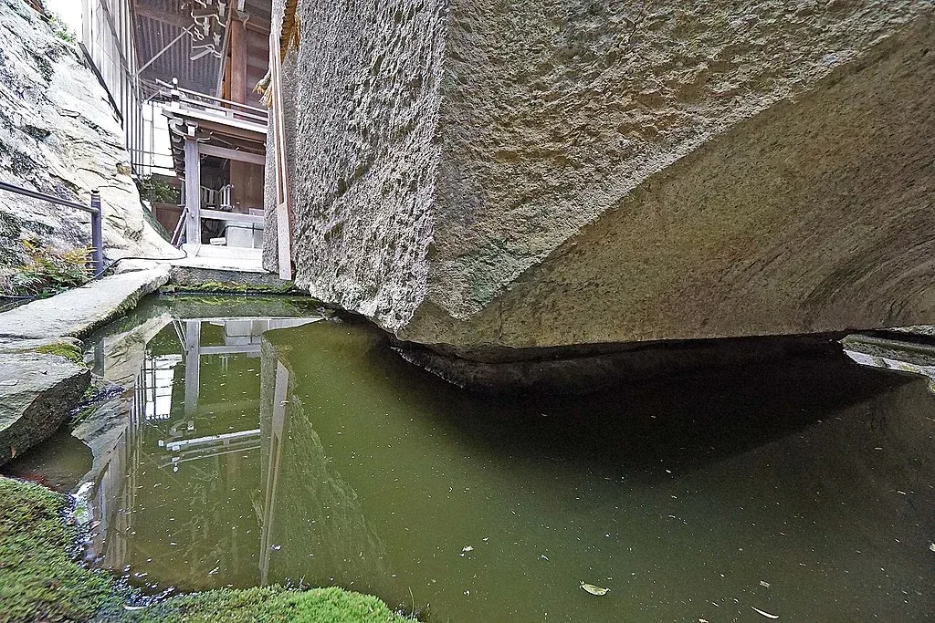 A close view of the Ishi no Hoden megalith with precisely cut edges rising above a pool of still water, its reflection and the sky mirrored in the calm surface
