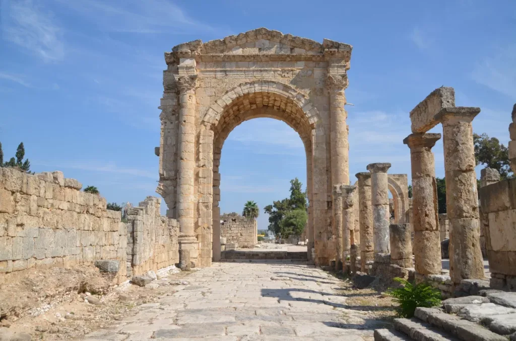 Ancient Roman triumphal arch and paved colonnaded street at the Baalbek temple complex in Lebanon, surrounded by weathered ruins under a bright blue sky