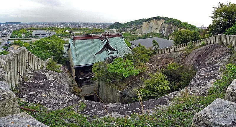 A top-down view of the Tatsuyama quarry historic site, showing a traditional Japanese building with a blue-green roof, the modern city, and steep greenery-covered quarry walls in the distance