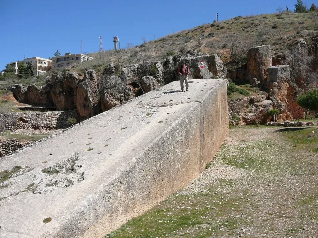 A man standing atop a colossal limestone monolith in the Baalbek quarry, next to a small Lebanese flag, emphasizing the monument's immense scale with hills and buildings in the distance