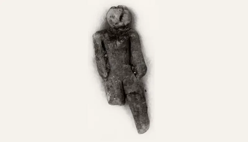 The Nampa figurine: a small, dark anthropomorphic clay doll with a round head, a simply shaped torso, arms at its sides, and elongated legs, shown against a light background.