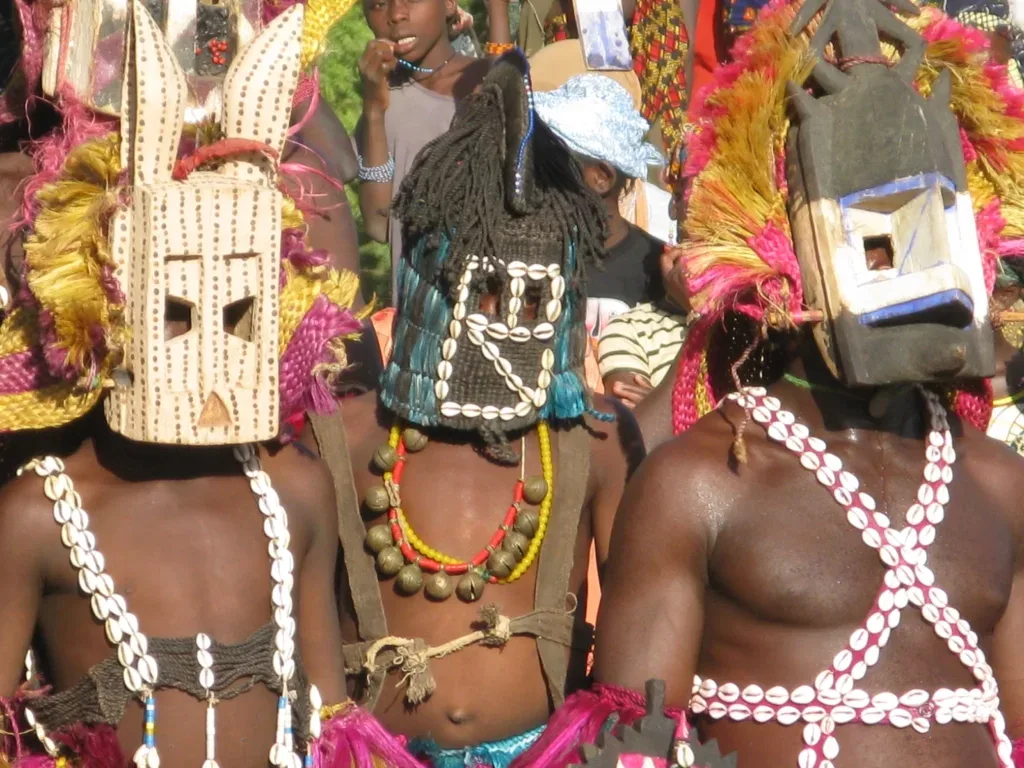 Three Dogon dancers in carved wooden masks and skirts of dyed leaves, with cowrie shell necklaces, performing before a gathered crowd