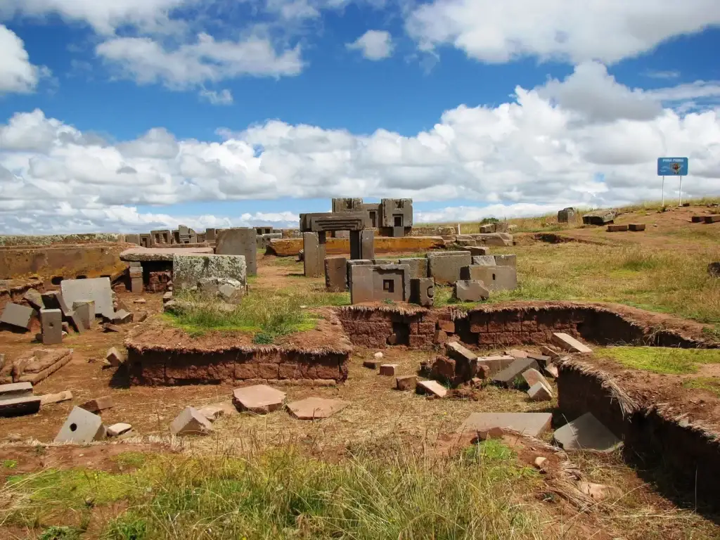 Wide panoramic view of the Puma Punku archaeological site with scattered megalithic ruins and geometrically shaped stone blocks under a blue sky